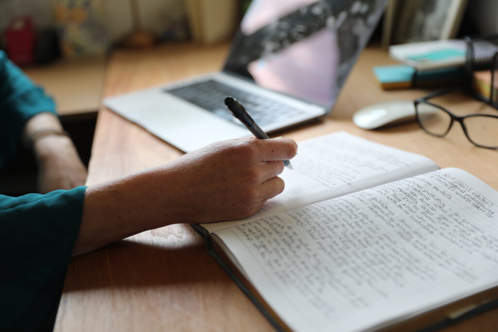 A notebook with handwritten notes in it and a pen. A laptop in the background